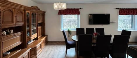 Un coin repas comprenant un buffet en bois, un lustre, un téléviseur fixé au mur et une fenêtre donnant sur des arbres.