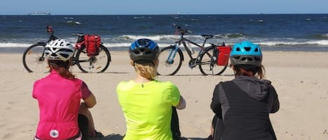 Trois personnes sont assises sur une plage de sable, avec des vélos et des casques, et elles font face à l’océan.