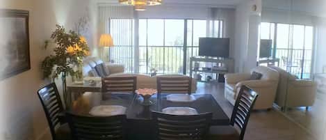 Een eetruimte met een tafel, stoelen, een kroonluchter, een bank, een televisie en een balkon met gordijnen.