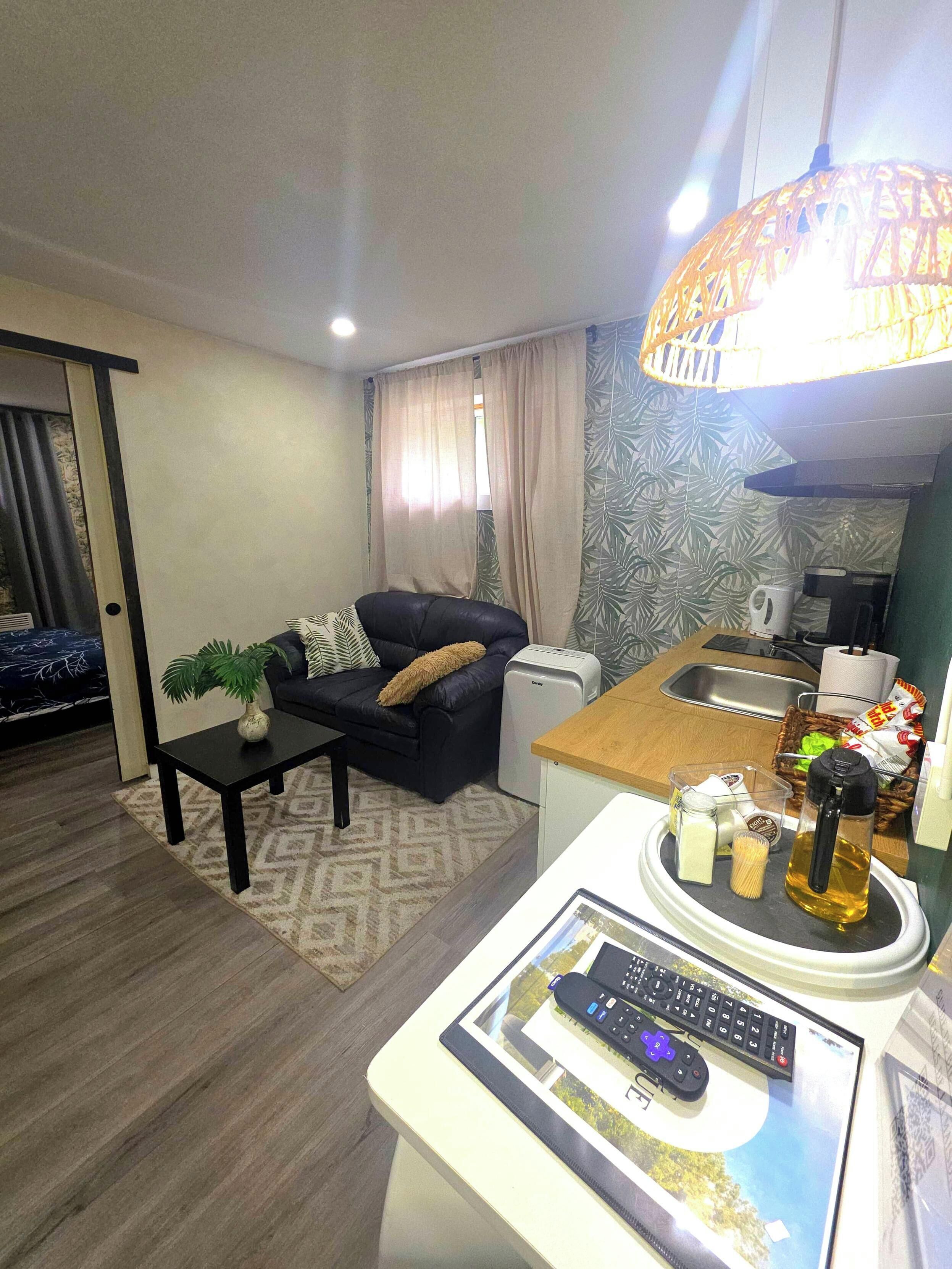 A living room with a sofa, coffee table, and a small kitchen area with a sink and a kettle.