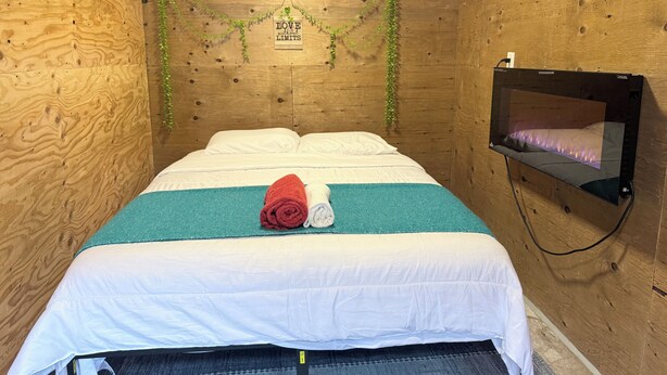 Une petite pièce chaleureuse avec un mur en bois, un lit avec une literie blanche et turquoise, une serviette rouge et un téléviseur fixé au mur.