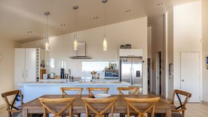 A modern kitchen with a central island, wooden chairs, pendant lighting, and a refrigerator. - Miro Ridge in Palm Beach (Waiheke Island)