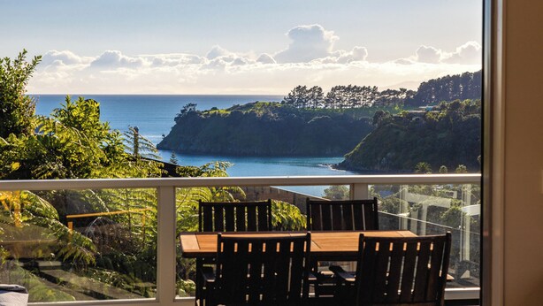 A balcony with a table and chairs, overlooking a coastal landscape with trees and a view of the sea. - Miro Ridge in Palm Beach (Waiheke Island)