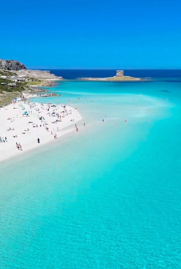 Ein Strand mit klarem türkisblauem Wasser, einem sandigen Ufer und einer kleinen Turmstruktur in der Ferne.