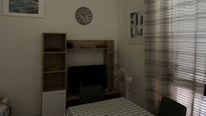 Un dormitorio con cama, escritorio, silla, televisor, reloj y ventana con cortinas.