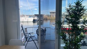 Una habitación con una puerta corrediza de vidrio que da a un balcón con vistas a barcos y una planta.