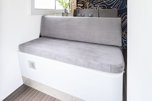 A grey upholstered bench with a white base, a window above, and a patterned wall to the right.