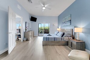 Ein Schlafzimmer mit Bett, Nachttischen, Deckenventilator, an der Wand montiertem Fernseher und Fenster mit Blick auf Grünflächen.