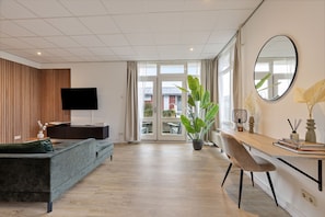 Un salon moderne avec un canapé, un téléviseur, un bureau avec une chaise, une plante en pot et une grande fenêtre.