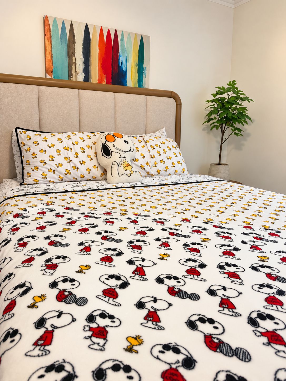 Ein Schlafzimmer mit einem Bett, das mit Snoopy-Bettwäsche, einem Snoopy-Kissen und einem farbenfrohen, abstrakten Gemälde über dem Bett ausgestattet ist.