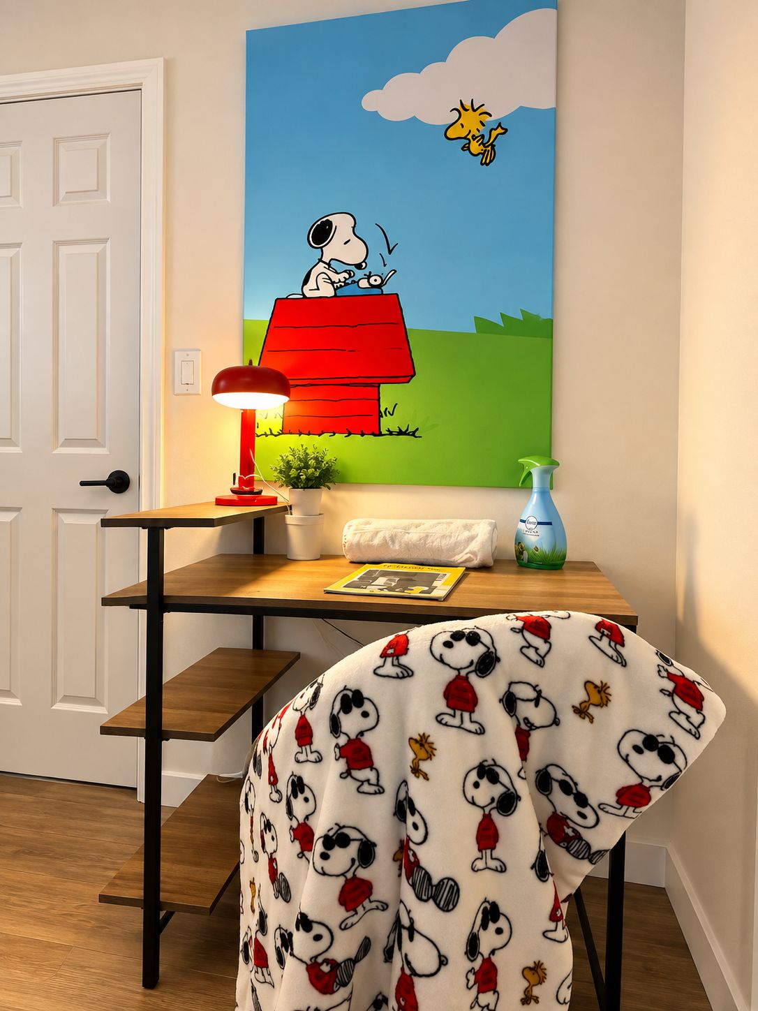 Quarto com pintura de Snoopy na parede, retratando-o em uma casa vermelha, uma escrivaninha com abajur, uma cadeira com tecido temático do Snoopy e uma porta.