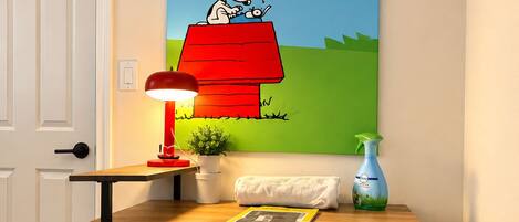 Une pièce avec un tableau représentant Snoopy accroché au mur, le tableau représentant Snoopy devant une maison rouge. On y trouve également un bureau avec une lampe, une chaise recouverte d’un tissu à motifs Snoopy et une porte.