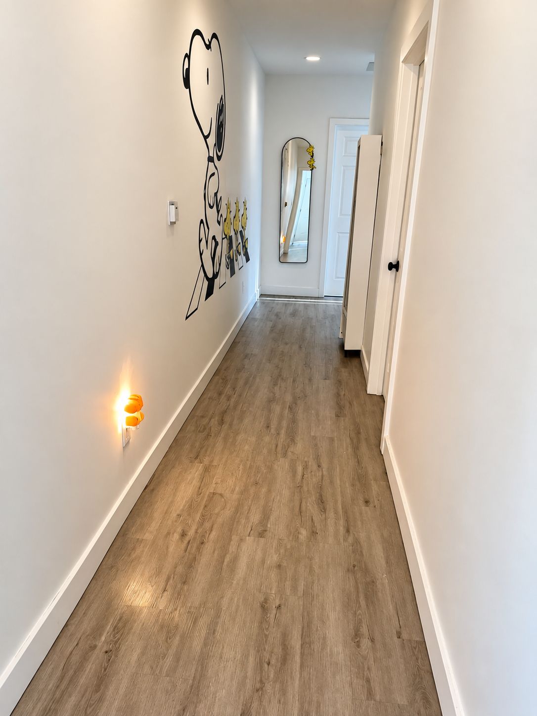 A hallway with a light-colored wooden floor, white walls, and a wall-mounted mirror.
