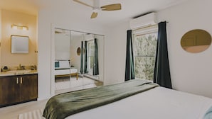 Un dormitorio con cama, espejo, ventilador de techo, ventana con cortinas y vista a zonas verdes.