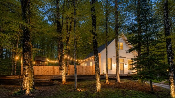 A white building with a wooden deck, surrounded by tall trees and string lights. - Schoolhouse | Fenced Yard | Family & Dog Friendly (Traverse City)