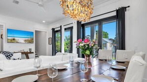 A dining area with a wooden table set for four, white chairs, a vase of flowers, and a painting of a beach scene on the wall. - 🦩Book Me Baby! Sun Of A Beach in Santa Rosa Beach~Golf Cart Included🦩 (Santa Rosa Beach)