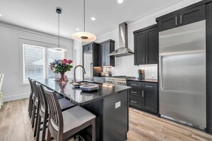 Une cuisine moderne dotée d’un îlot central, de meubles de couleur sombre, d’appareils électroménagers en acier inoxydable et d’un luminaire suspendu.