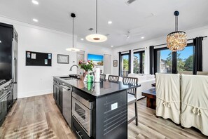 Een moderne keuken met een centraal eiland, roestvrijstalen apparatuur, hangende lampen en uitzicht op de buitenlucht door grote ramen.