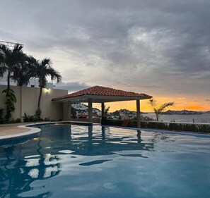 A swimming pool with a covered area, palm trees, and a view of a distant cityscape during sunset. - HOUSE LAS PALMAS ACAPULCO (Acapulco de Juárez)