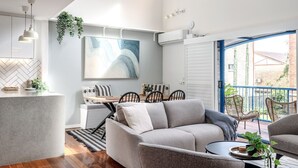 A modern living room with a sofa, dining table, and chairs, a kitchen area with a counter, and a balcony with outdoor seating. - Biggera Breeze Retreat (Biggera Waters)