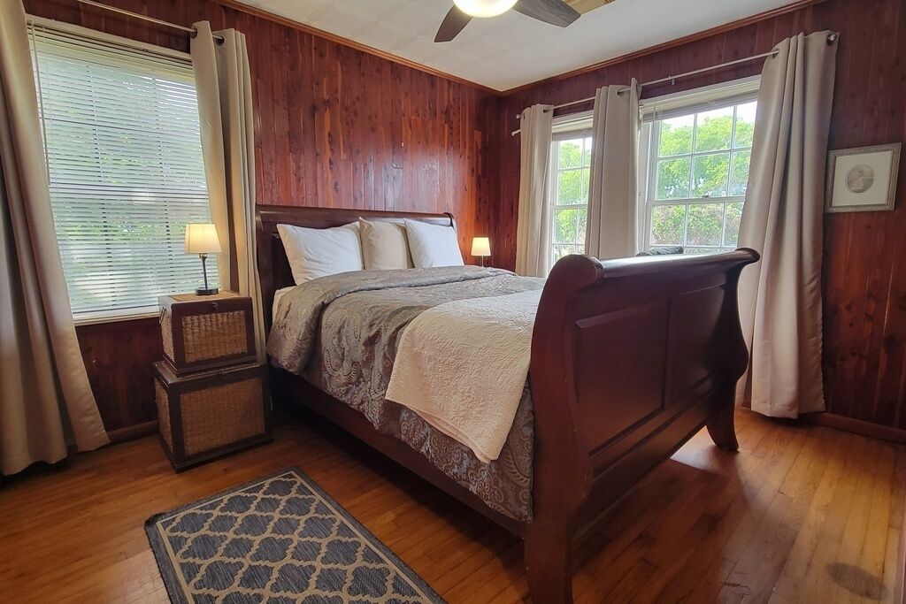 A wooden-paneled bedroom with a bed, bedside lamps, a window with curtains, and a small rug.
