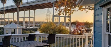 Um terraço com mesa e cadeiras, com vista para um corpo d'água onde se vê uma ponte e fios de eletricidade ao fundo.