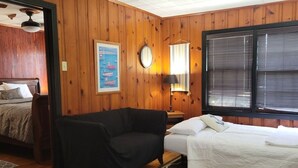 Una habitación con paredes de madera, una cama con ropa de cama blanca, un sofá negro, una mesita de noche con una lámpara, un cuadro enmarcado en la pared y una ventana con persianas.