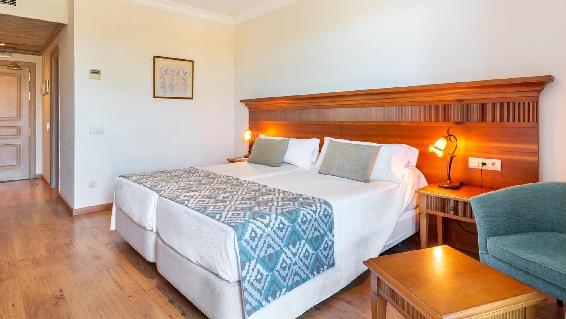 A hotel room with a double bed, bedside lamps, a small table, a chair, and a wooden headboard.