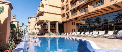 Zona de piscina con sillas de descanso, una piscina azul cristalina y un edificio de varios pisos con balcones.