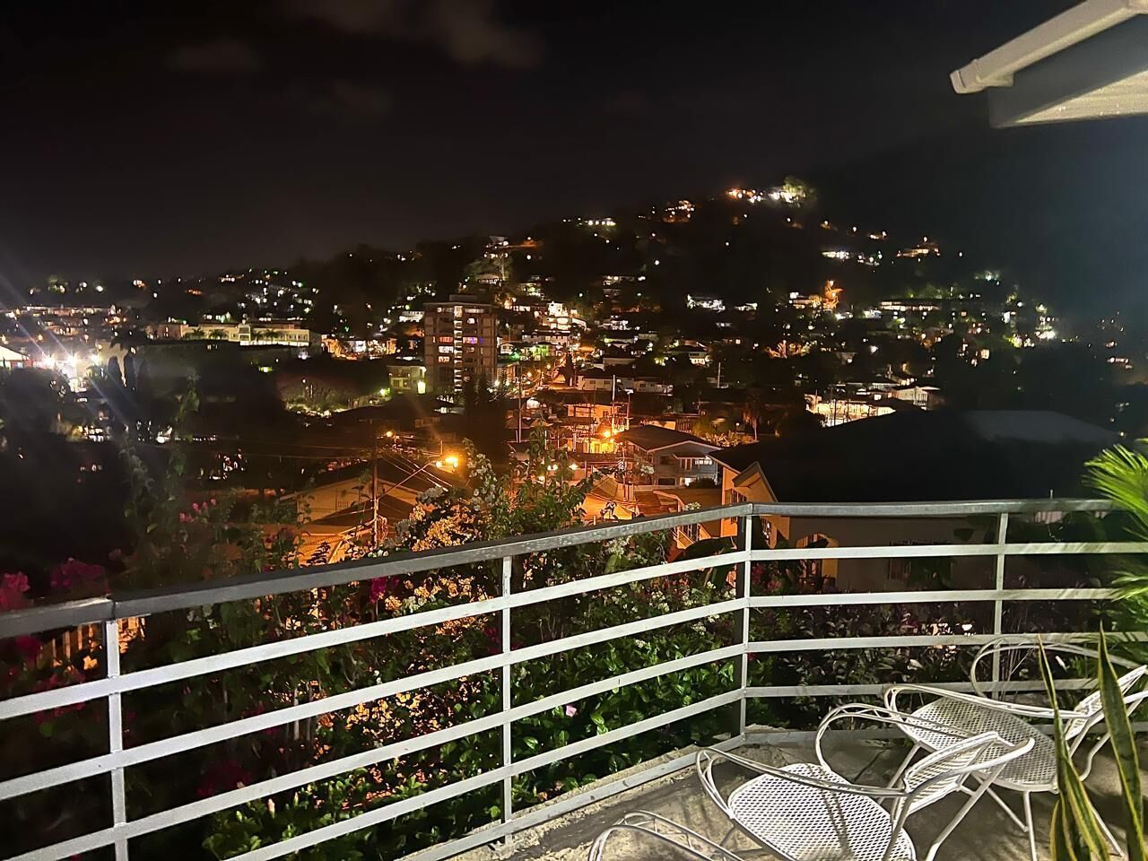 Une vue nocturne d’une ville côtière, avec des bâtiments illuminés et un balcon agrémenté de chaises.