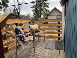 A wooden deck with a metal chair and a bench, a blue door, and a view of a house in the background. - Upper Pine Vacation Rental | Bend Living (Bend)