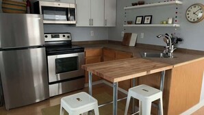 Een keuken met roestvrijstalen apparatuur, houten aanrechtbladen, witte kastjes, een houten kookeiland met krukken en een klok aan de muur.
