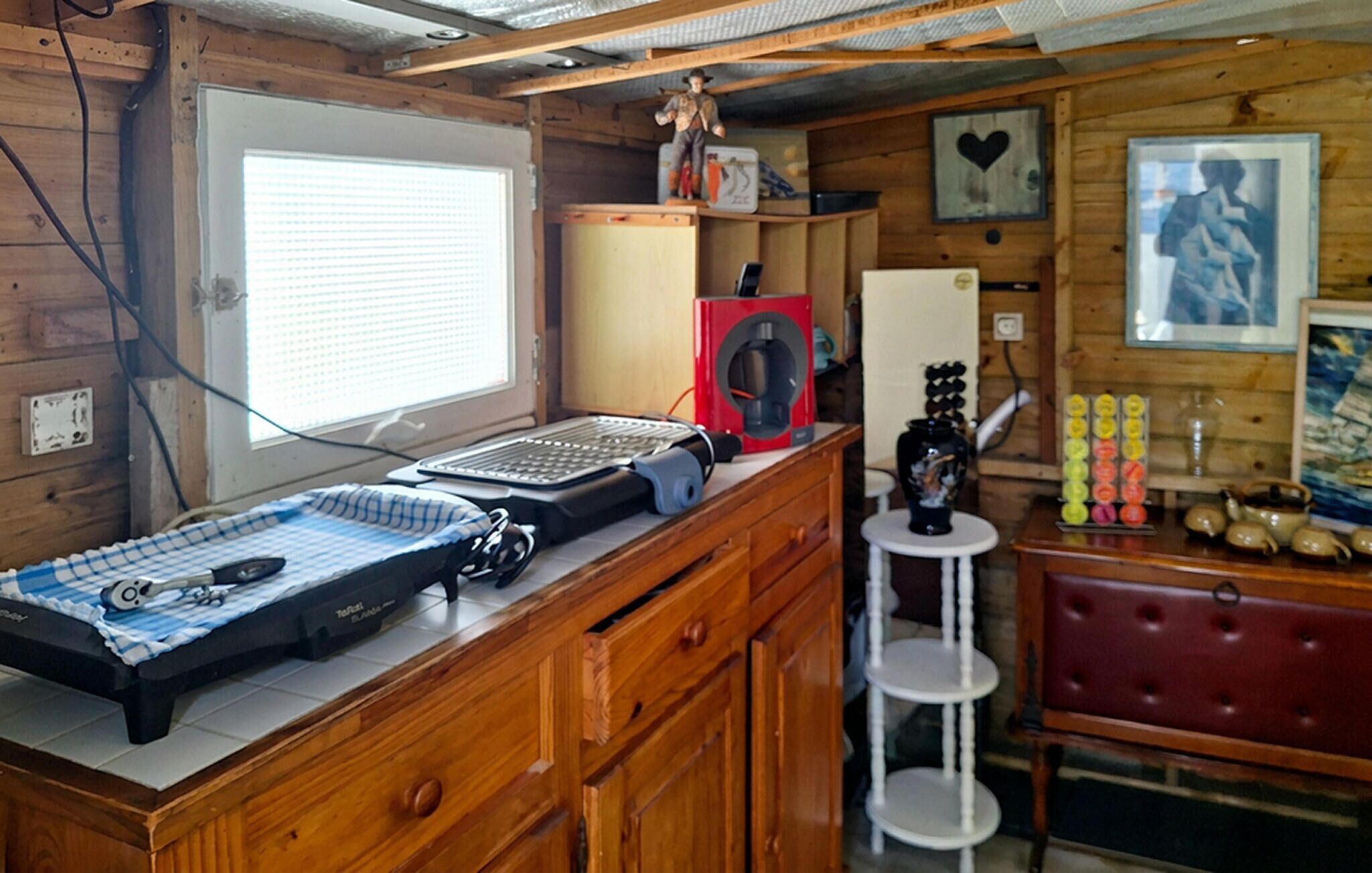 Une pièce de taille modeste, comprenant un bureau en bois, un ordinateur, une imprimante, une petite étagère avec des objets décoratifs et une fenêtre équipée de stores.