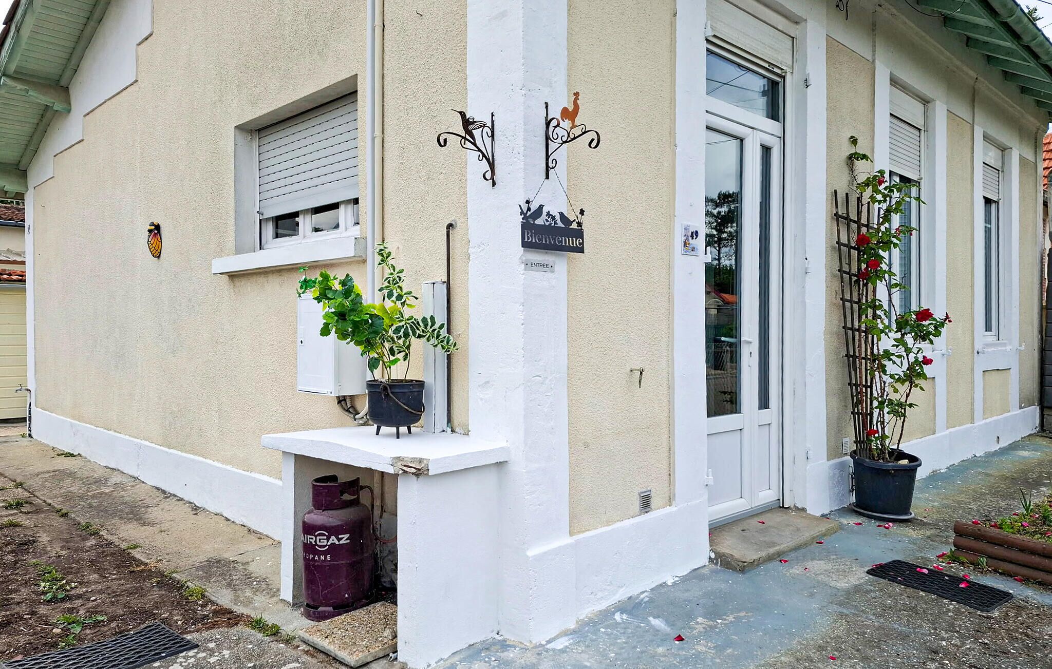 A building with a white facade, a 'Bienvenue' sign, a window with a shutter, and potted plants on the ground level.