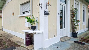 Een gebouw met een witte gevel, een 'Bienvenue' bord, een raam met luik en potplanten op de begane grond.
