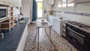 Een keuken met een betegelde spatwand, een fornuis, een spoelbak en een eethoek met een tafel en stoelen.