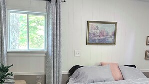 Ein Schlafzimmer mit Bett, einem Fenster mit Vorhängen, einem Nachttisch mit Lampe, einem Schminktisch und einem Bild an der Wand.