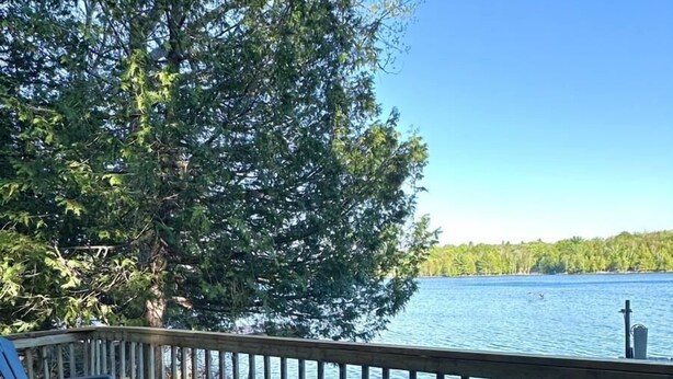 Eine Holzterrasse mit Adirondack-Stühlen, mit Blick auf einen ruhigen See mit Bäumen im Hintergrund.