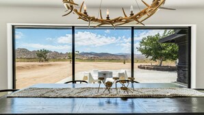 A dining area with a view of a desert landscape through large windows, a wooden table, and a unique antler chandelier.