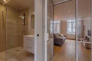 Un baño moderno con cabina de ducha de vidrio, tocador blanco y puerta de vidrio que da a una sala de estar con sofá y escritorio.
