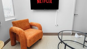Huoneessa on seinälle asennettu televisio, jossa näkyy teksti »NETFLIX», pehmeä oranssi nojatuoli, lasinen pöytä, jonka päällä on valkoinen lintuhahmo, sekä ikkuna, josta avautuu näkymä rakennuksiin.