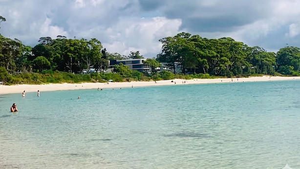 Una playa con aguas transparentes, una orilla arenosa y un edificio de varios pisos al fondo.