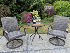 Zwei Korbstühle mit Metallgestellen und ein kleiner runder Tisch mit einer Flasche und Gläsern.