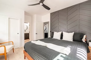 Een slaapkamer met een bed, een plafondventilator, een nachtkastje, een stoel en een badkamer met een spiegel.