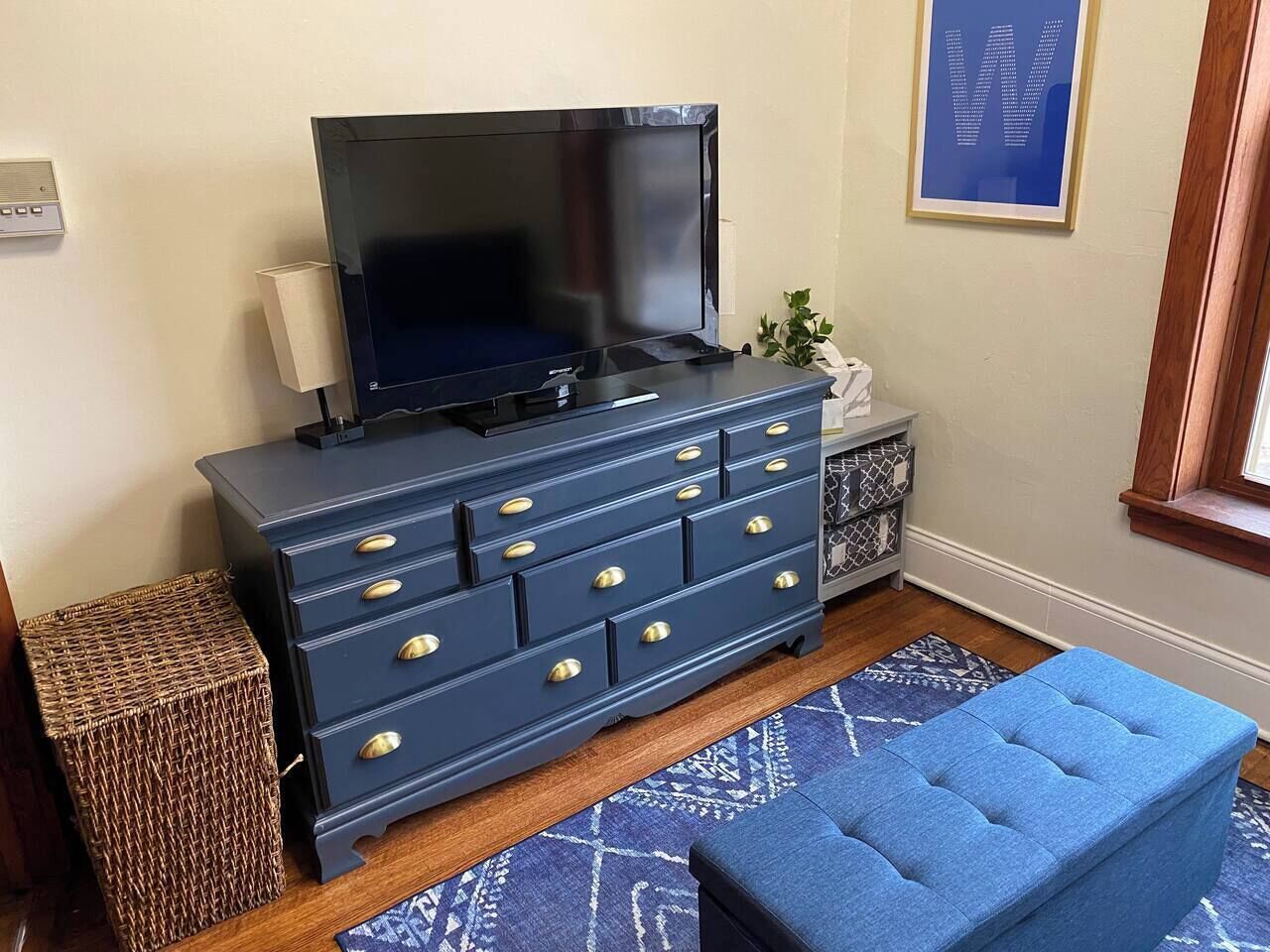 Une pièce avec une commode bleu foncé, un téléviseur posé dessus, un pouf bleu, un tapis à motifs et une petite plante.