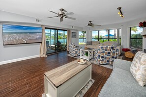 Ein geräumiges Wohnzimmer mit großem Fernseher, zwei Deckenventilatoren, einer Couch, einem Couchtisch und Meerblick durch große Fenster.