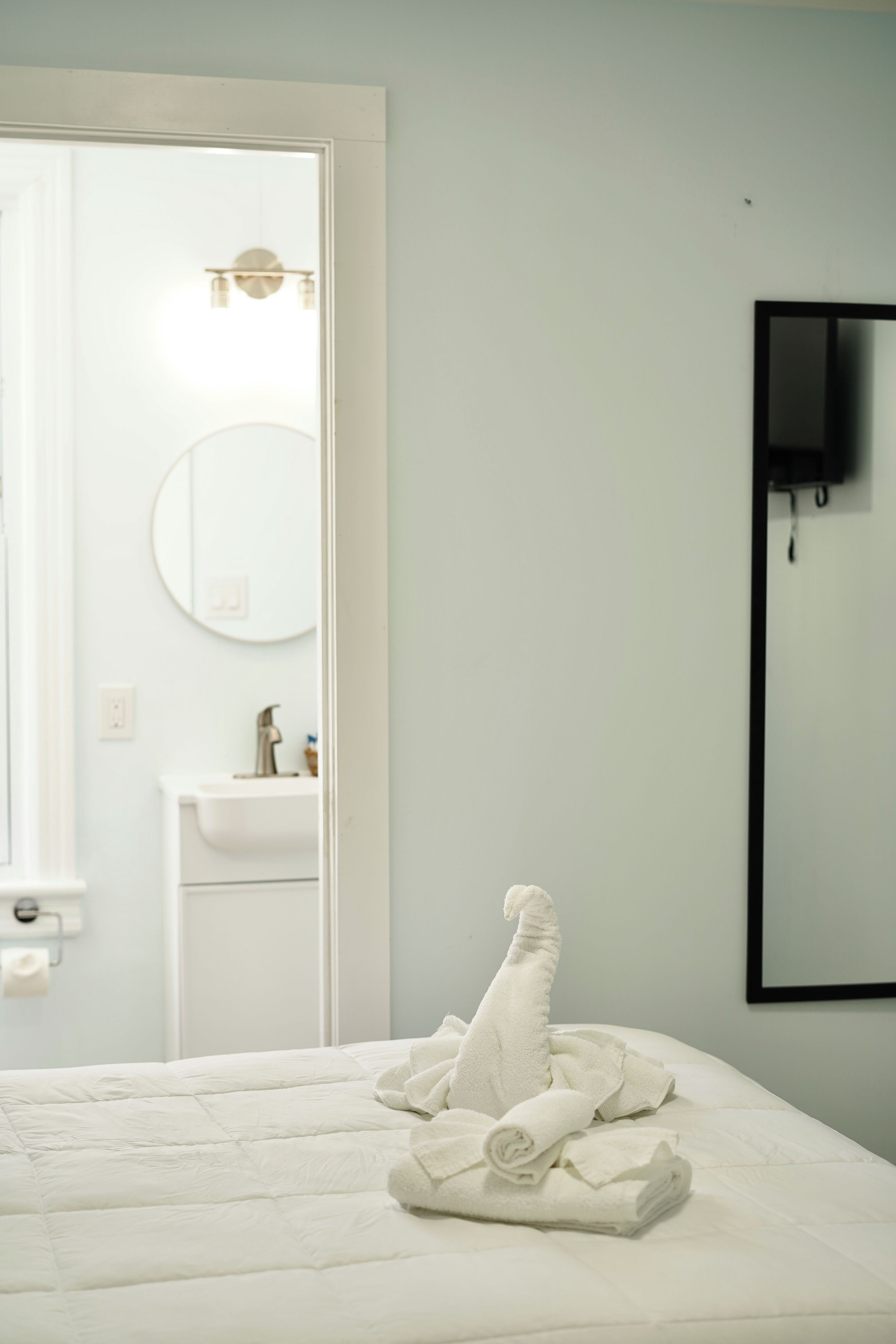 Une chambre à coucher avec un lit, une serviette pliée, un téléviseur fixé au mur et une salle de bain visible à travers une porte ouverte.