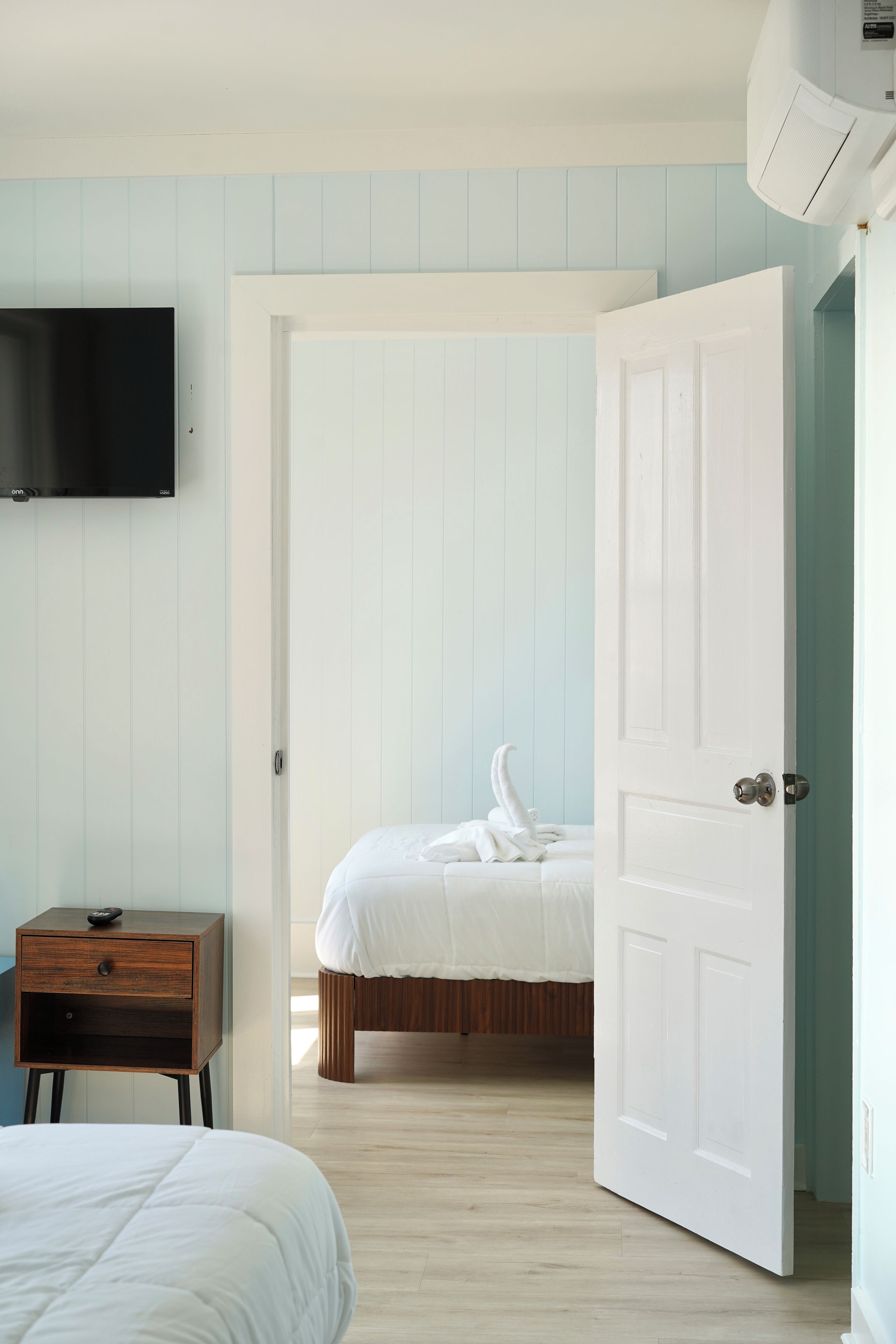 A bedroom with a bed, a nightstand, a television mounted on the wall, and a door leading to another room.