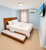 Un dormitorio con cama de madera, un banco, una lámpara en el techo, un equipo de aire acondicionado y un televisor montado en la pared.