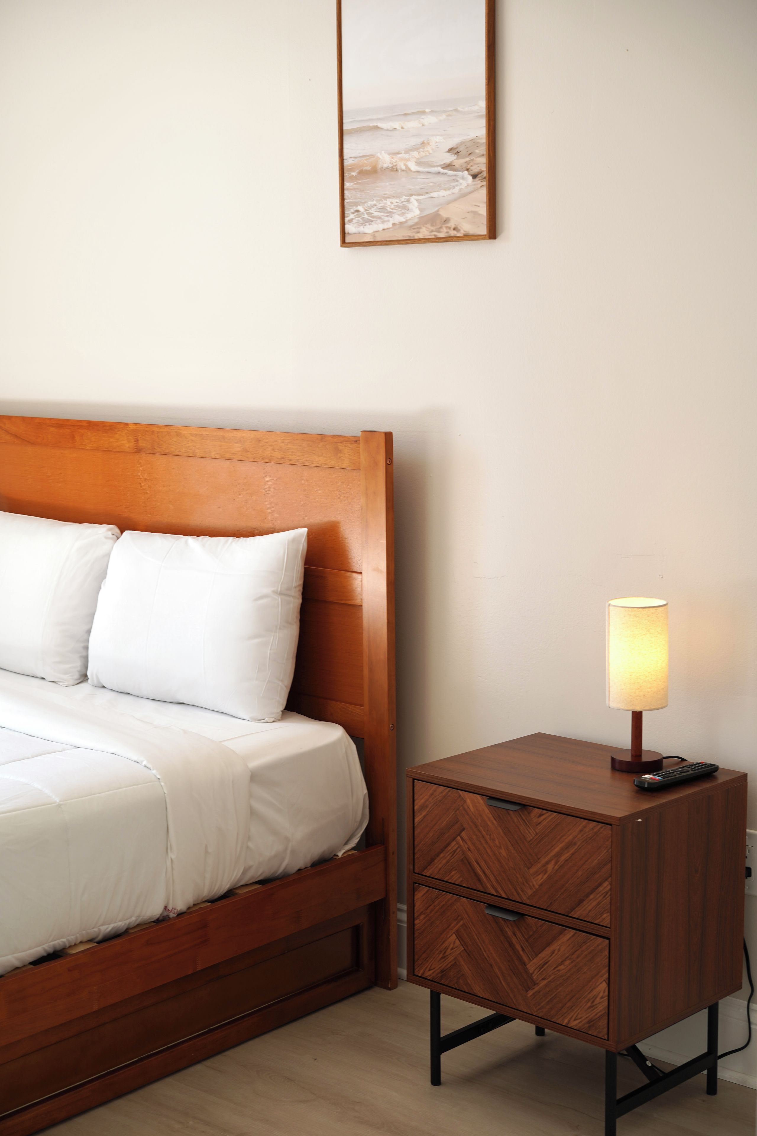 Una cama bien tendida con sábanas blancas, un cabecero de madera y una mesita de noche con cajones y una lámpara.